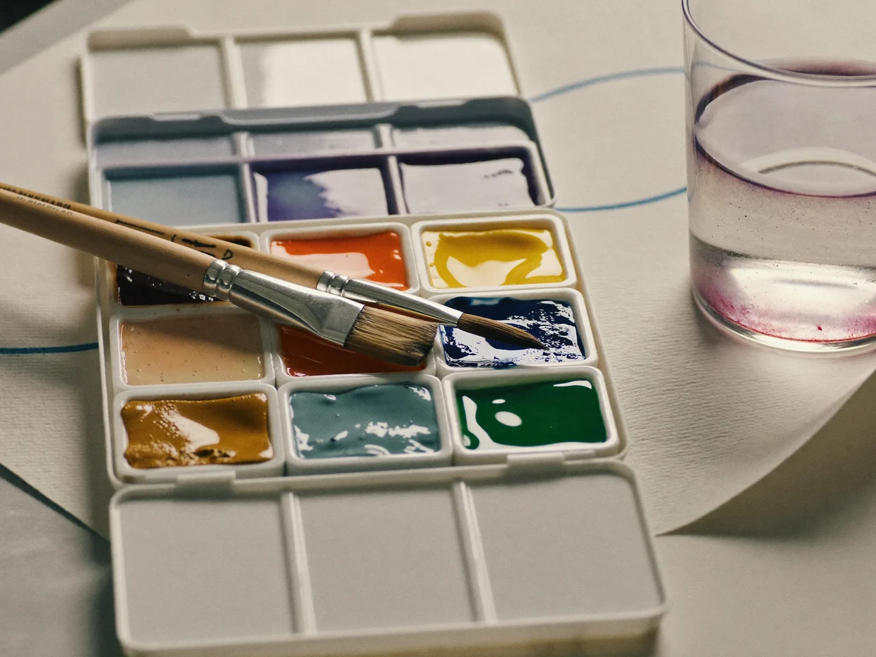 Aquarellfarbkasten mit geoeffneten Napfchen, nassen Pinseln und Wasserglas auf Papier